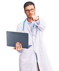 Handsome young man with bear wearing doctor uniform working using computer laptop pointing with finger to the camera and to you, confident gesture looking serious