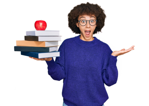 Young hispanic girl wearing glasses holding books and red apple celebrating victory with happy smile and winner expression with raised hands