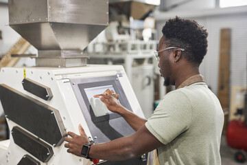 Back view of Black young man using touch screen control panel while operating machines in food factory workshop, copy space - obrazy, fototapety, plakaty