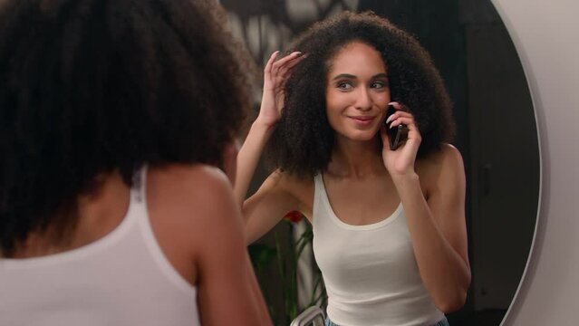 Happy African woman girl talking mobile phone laughing talk speaking smartphone call friend pleasant conversation looking at bathroom mirror check hair checking fix hairdo hairstyle laugh gossiping