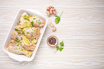 Raw uncooked chicken legs with seasonings, herbs, lemon in white ceramic casserole top view on light wooden rustic background. Preparing healthy meal with marinated chicken drumsticks. Space for text.
