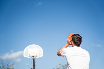 Young Caucasian boy with his back turned, wearing a white t-shirt throwing a basketball at a metal...