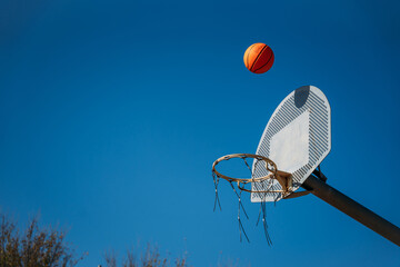 Basketball basket seen from below and one side while an orange ball is falling from above. Contrasted with a blue sky on a sunny, clear day.