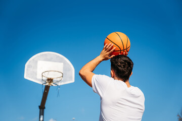 Young Caucasian boy with his back turned, wearing a white t-shirt ready to throw a basketball at a metal basket and the clear blue sky on a sunny day.