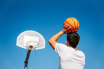 Young Caucasian boy with his back turned, wearing a white t-shirt ready to throw a basketball at a metal basket and the clear blue sky on a sunny day.
