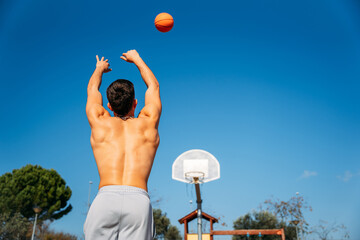 Young Caucasian guy seen from behind, muscular shirtless while throwing a basketball at a metal basketball court on a sunny day with clear blue sky.