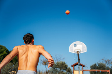 Young Caucasian guy seen from behind, muscular shirtless while throwing a basketball at a metal basketball court on a sunny day with clear blue sky.