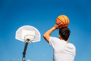 Young Caucasian boy with his back turned, wearing a white t-shirt ready to throw a basketball at a metal basket and the clear blue sky on a sunny day.