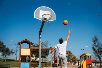 Chico joven cauc&aacute;sico de espaldas, con camiseta blanca saltando y ganastando una pelota de b&aacute;squetball a una ganasta de metal y el cielo azul despejado en un d&iacute;a soleado.