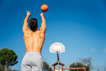 Young Caucasian guy seen from behind, muscular shirtless while throwing a basketball at a metal basketball court on a sunny day with clear blue sky.