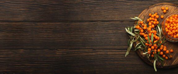 Sea buckthorn ripe berries in glass jar and branches with leaves top view on dark wooden rustic background, great for skin, heart, vessels and immune system. Copy space..