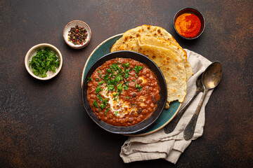 Traditional Indian Punjabi dish Dal makhani with lentils and beans in black bowl served with naan flat bread, fresh cilantro and two spoons on brown concrete rustic table top view. - obrazy, fototapety, plakaty