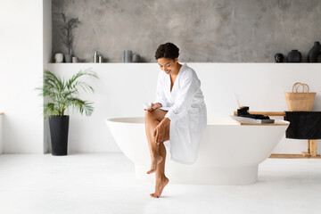 Beautiful black female applying cream on legs while sitting in luxury bathroom