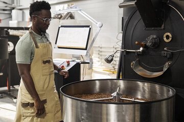 Portrait of African American young man watching coffee roasting process and operating machines,...