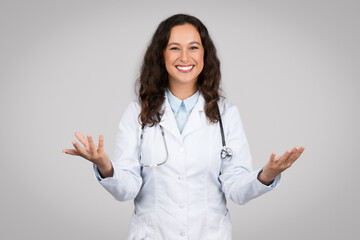 Portrait of cheerful european female doctor in workwear gesturing and talking at camera on grey...