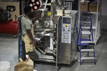Back view of Black male worker setting up packaging machine at small coffee production factory,...