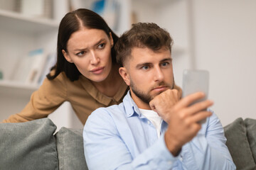 Jealous wife peeking over husband's shoulder as he texts indoors