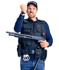 Young handsome man wearing police uniform holding shotgun annoyed and frustrated shouting with...