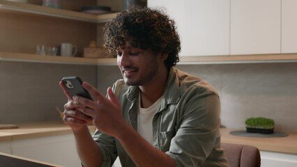 Moving shot carefree happy Indian man browsing smartphone laugh Arabian male smiling scrolling social media network app looking at mobile phone laughing smile enjoy funny video online at home kitchen