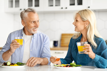 Happy Senior Spouses Chatting And Drinking Orange Juice During Breakfast In Kitchen