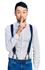 Hispanic man with beard wearing hipster look with bow tie and suspenders asking to be quiet with finger on lips. silence and secret concept.