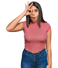Young hispanic girl wearing casual t shirt doing ok gesture shocked with surprised face, eye looking through fingers. unbelieving expression.