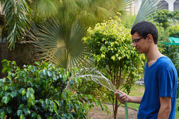 Young man watering garden plants using a water pipe at home. A person, tall asian teenage boy dusts...