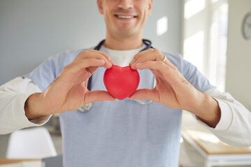 Close up of smiling friendly male doctor cardiologist holding red heart in his hands in clinic and...