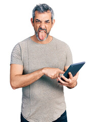 Middle age hispanic with grey hair using touchpad device sticking tongue out happy with funny expression.