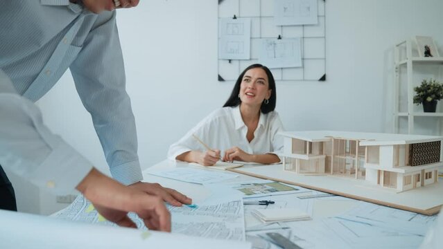 Skilled caucasian interior designer working with house model while discussing with asian architect engineer about house plan on meeting table with blueprint and house model scatter around. Immaculate.