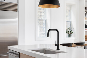 A kitchen sink detail with a black faucet under a black and gold pendant light, white oak island, white countertop, and decorations in the background.	
