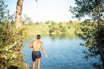 Unrecognizable muscular young guy seen from behind thinking about jumping into water from a rope/zip line. Caucasian boy doing water sport.