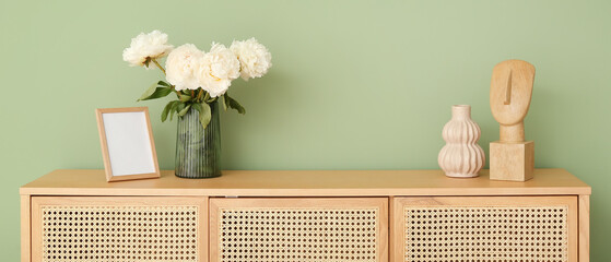 Vase with white peonies and decor on wooden dresser near green wall in room