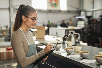 Side view portrait of smiling young woman taking notes on clipboard during cupping and quality...