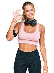 Young blonde woman wearing gym clothes and using headphones smiling positive doing ok sign with hand and fingers. successful expression.