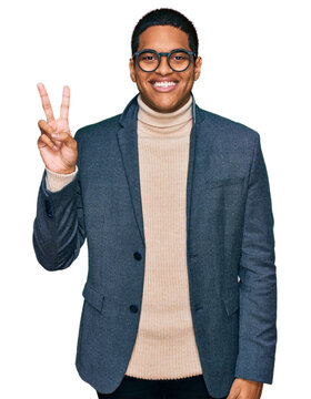 Young handsome hispanic man wearing business jacket and glasses smiling with happy face winking at the camera doing victory sign. number two.