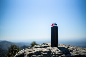 Black unbranded canteen or sports bottle on a very sunny and hot summer day resting on a pile of stones on a mountain with a very blue sky in the background.
