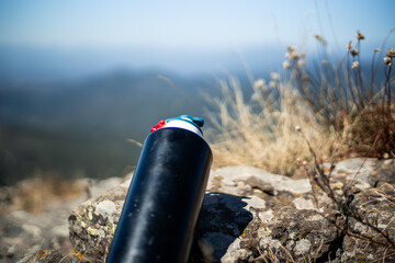 Black unbranded canteen or sports bottle on a very sunny and hot summer day resting on a pile of stones on a mountain.