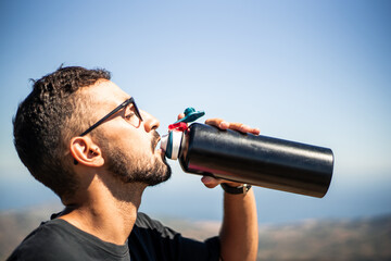 Young Caucasian man of North African nationality thirstily drinking water from a black unbranded canteen or bottle on a very sunny and hot summer day. Profile view.
