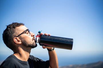 Young Caucasian man of North African nationality thirstily drinking water from a black unbranded canteen or bottle on a very sunny and hot summer day. Profile view.