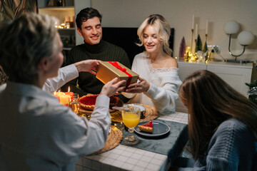 Pretty happy young woman receiving gift from loving mother-in-law sitting with family at dinner feast table at home on Christmas Eve. Blonde female giving present to beloved daughter on holidays.