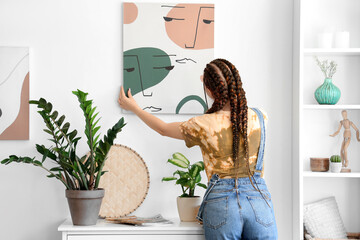 Young woman with dreadlocks hanging painting on wall at home, back view