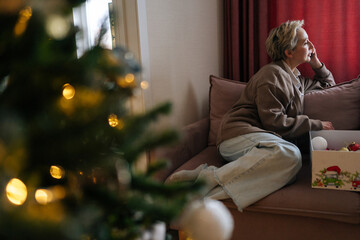 Remote view of happy gray-haired woman talking smartphone and choosing festive xmas toys sitting on sofa in living room. Preparation for holidays, decorates Christmas tree for New Year at home.