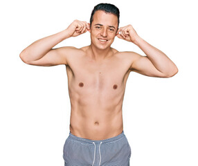 Fototapeta premium Handsome young man wearing swimwear shirtless smiling pulling ears with fingers, funny gesture. audition problem