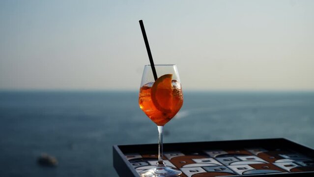 Closeup of glass with Aperol Spritz (aperitif cocktail consisting of prosecco, Aperol and soda water) on background of beautiful seascape on summer sunny day. Concept of vacations and travel.