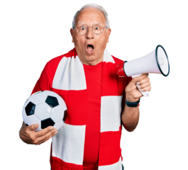 Senior man with grey hair football hooligan holding ball and using megaphone afraid and shocked with surprise and amazed expression, fear and excited face.