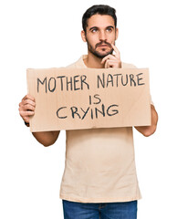 Young hispanic man holding mother nature is crying protest cardboard banner serious face thinking about question with hand on chin, thoughtful about confusing idea