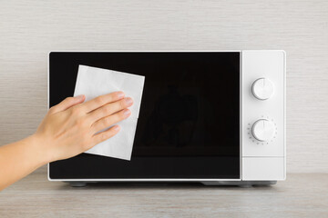 Young adult woman hand holding dry white paper napkin and wiping dark black glass door surface of white microwave oven on table top at home kitchen. Closeup. Regular cleanup. Front view. - obrazy, fototapety, plakaty