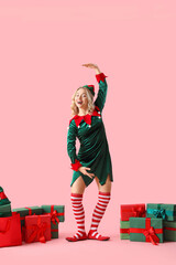 Funny young woman dressed as elf with Christmas gifts on pink background