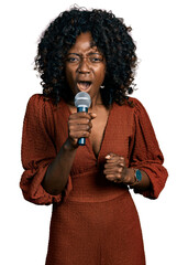 African woman with curly hair singing song using microphone angry and mad raising fist frustrated...